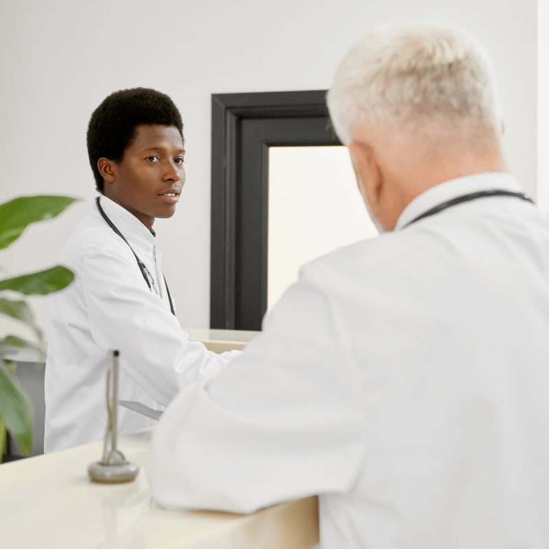 Selective focus of younf african doctor with stethoscope on neck standing behind reception and looking at coworker. Back view of incognito eldery therapist leaning on stand with plants.