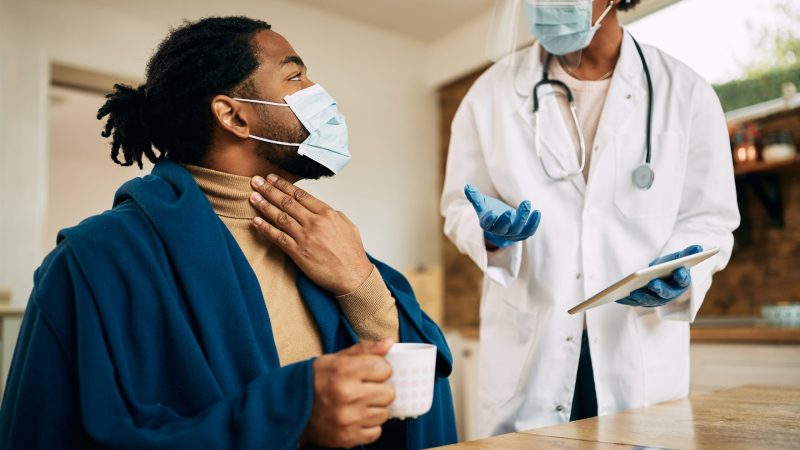 African American man wrapped in blanked talking to his doctor and complaining about sore throat during a home visit. Both of them are wearing face masks due to coronavirus pandemic.