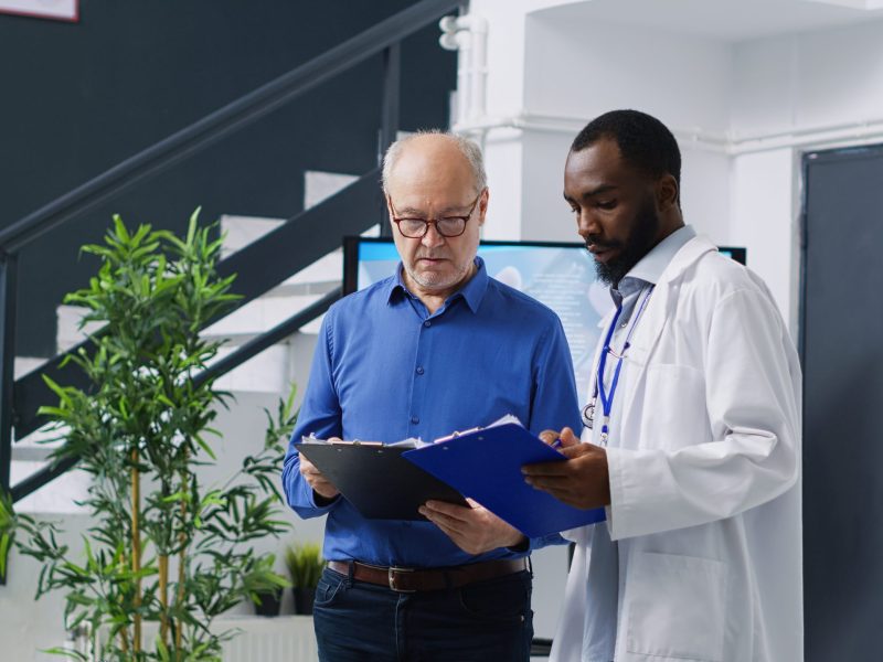 Specialist explaining disease expertise to elderly patient during checkup visit consultation in hospital waiting area. Senior man man filling medical insurance documents, medicine service